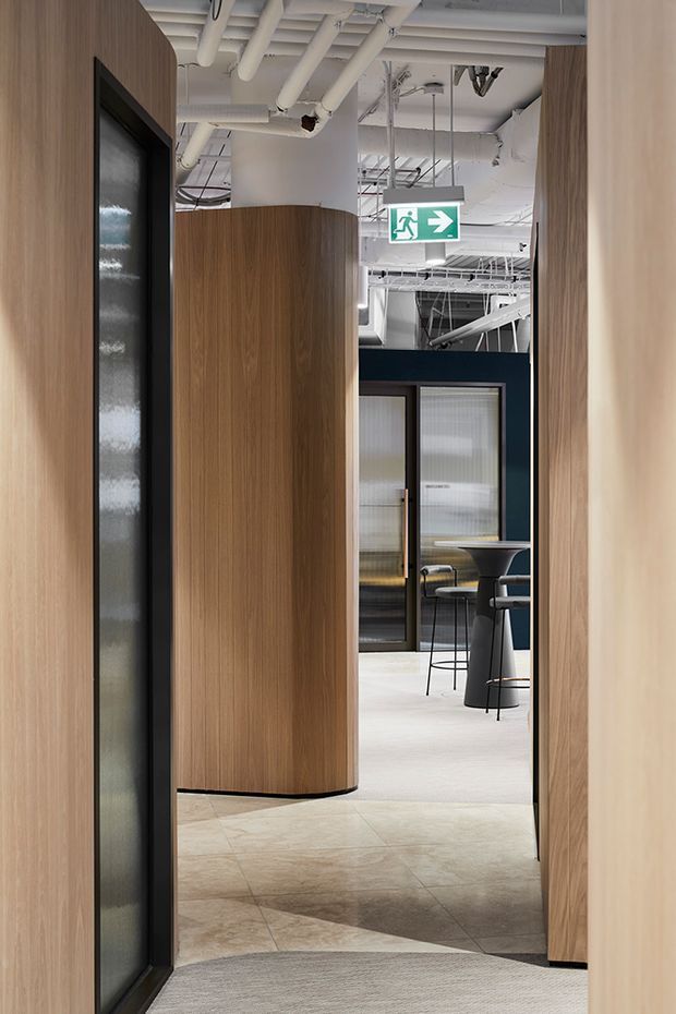 A contemporary commercial interior hallway leading into an open office or hospitality-style breakout area. The corridor is lined with curved timber-look wall panels and dark-framed translucent glass doors, while the exposed ceiling services, white pipes, suspended lighting, and emergency exit sign create an industrial office character. A small bar-height table and stools are visible in the adjoining space.