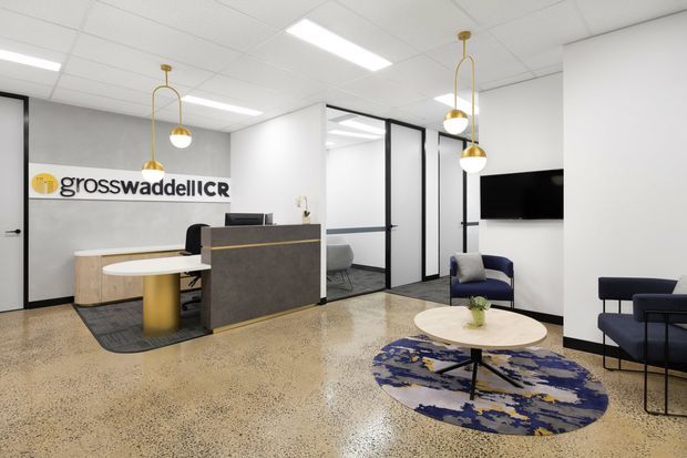 Bright contemporary commercial reception area with a polished terrazzo floor, white acoustic tile ceiling, glass-partitioned offices, and a dark grey reception desk. The space includes a wall-mounted Gross Waddell ICR sign, sculptural gold pendant lights, a round meeting table, patterned circular rug, and navy lounge seating.