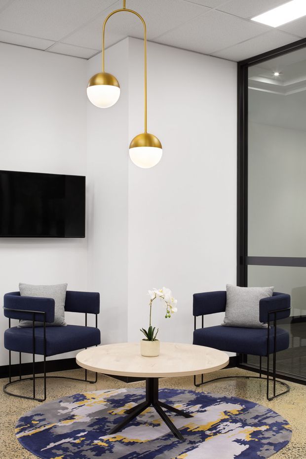 A contemporary commercial lounge or waiting area with two navy upholstered armchairs, a round pale wood coffee table, and a bold abstract circular rug. White walls and a suspended tiled ceiling are accented by sculptural brass-and-white globe pendant lights. A wall-mounted television and black-framed glass partition suggest an office reception or meeting environment.