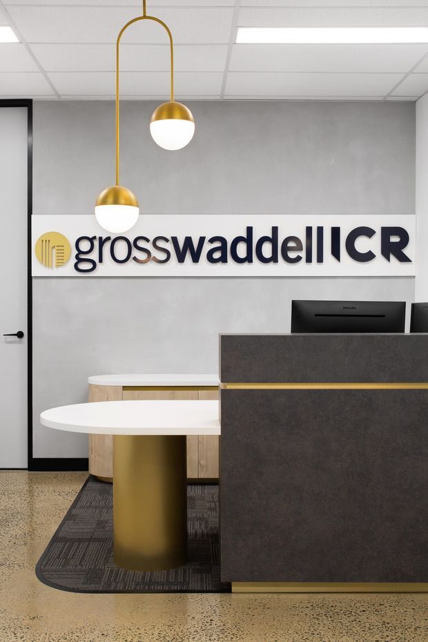 A modern commercial reception area featuring a dark stone-look reception desk with a slim brass-gold accent, a white curved visitor table, timber and brass cabinetry, terrazzo flooring, grey textured wall finish, and a large illuminated Grosswaddell CR sign. Two gold pendant globe lights hang from a suspended ceiling grid.