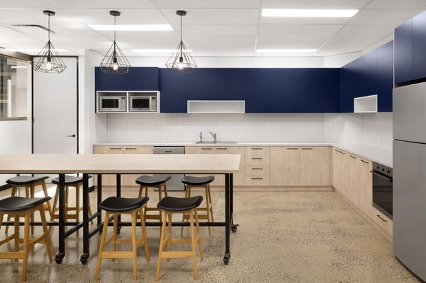 A contemporary commercial-style kitchen and communal dining area with pale timber cabinetry, a dark navy-blue feature wall and upper cabinets, white geometric tiled splashback, stainless-steel appliances, and a large mobile island table surrounded by timber stools. Suspended geometric pendant lights and a tiled acoustic ceiling complete the clean, functional fit-out.
