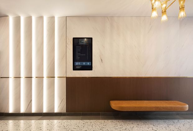 A refined contemporary lobby or entry area featuring a floating timber bench, dark wood wall panelling, large light veined stone wall panels, vertically illuminated wall fins, and a wall-mounted digital control or information panel. Decorative pendant lights hang above, and the polished terrazzo-like floor contributes to a luxurious, calm atmosphere.
