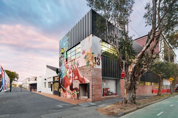 Street-level exterior view of a contemporary multi-storey commercial building with black vertical metal cladding, exposed red brick, extensive colourful mural artwork, and large glazed openings. The building sits beside a tree-lined urban street with a marked cycle lane and neighbouring low-rise commercial structures.