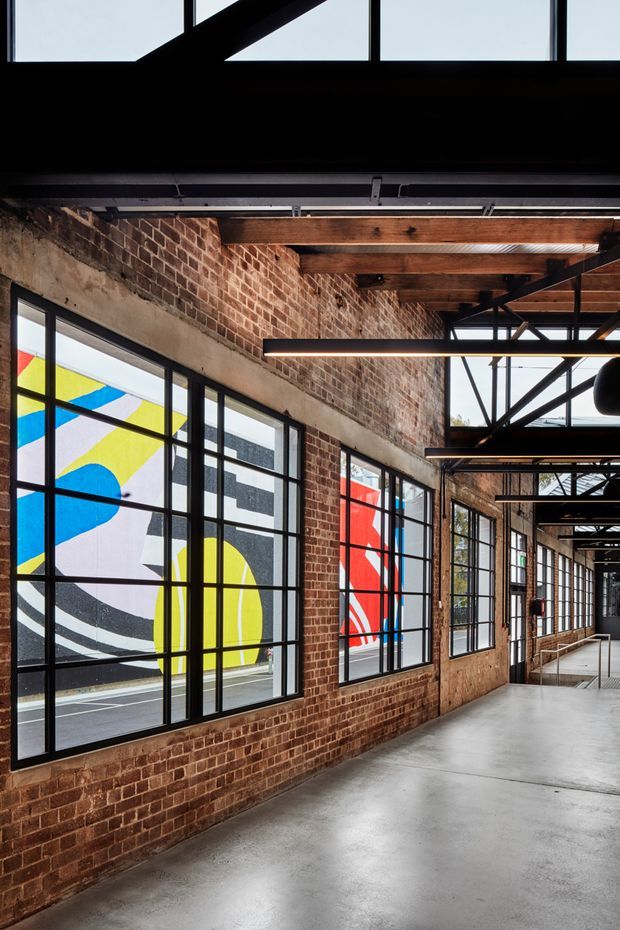 A long industrial-style interior corridor or circulation space with exposed red brick walls, polished concrete flooring, black-framed industrial windows, exposed timber ceiling beams, and visible steel structural bracing. Large colourful geometric artwork is visible through the glazing, adding bold red, blue, yellow, black, and white accents.