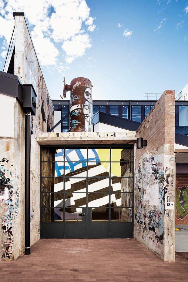 Exterior view of an adaptive-reuse style urban building entrance, featuring a weathered concrete and graffiti-covered facade, black metal-framed glazed industrial doors, exposed brick paving, and a prominent rusted cylindrical tank above the entry. Modern dark-framed windows and roof forms are visible behind the retained industrial fabric.