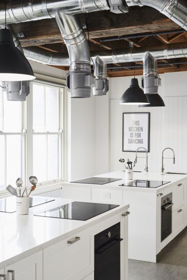 Bright industrial-style kitchen with white flat-panel cabinetry, expansive white benchtops, integrated black appliances, twin sinks and tall gooseneck taps. Exposed timber ceiling structure, extensive silver ductwork, and black pendant lights create a strong industrial character, while large windows provide abundant natural light.