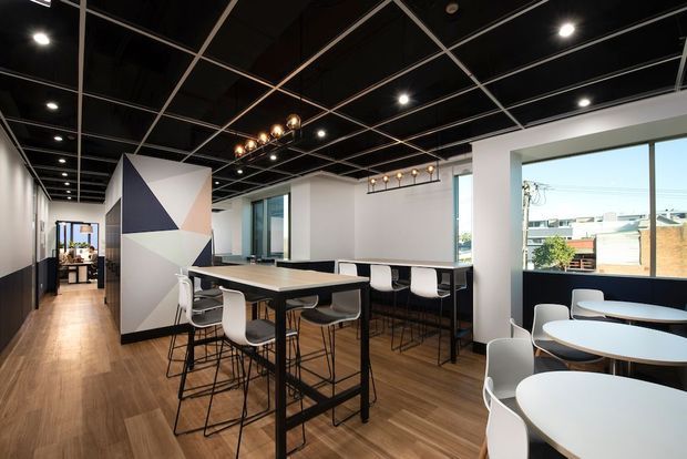 An open-plan contemporary commercial workspace or office interior with warm timber flooring, white walls, black-framed glazing and a dramatic black exposed grid ceiling. The space contains communal high tables with stools, round café-style tables, white task chairs, and a geometric navy, blush and white feature wall. Large windows overlook an urban streetscape, while pendant and recessed lighting provide illumination.