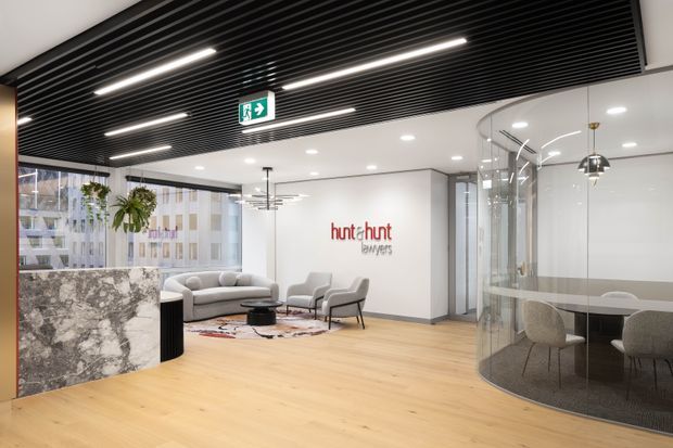 A contemporary commercial office reception and breakout area with pale timber flooring, a marble-patterned reception counter, soft seating, indoor plants, and a Hunt & Hunt Lawyers logo on the wall. A curved glass meeting room with a round table is visible on the right. The space features a black linear ceiling grille with integrated strip lighting, white walls, and large windows overlooking an urban setting.