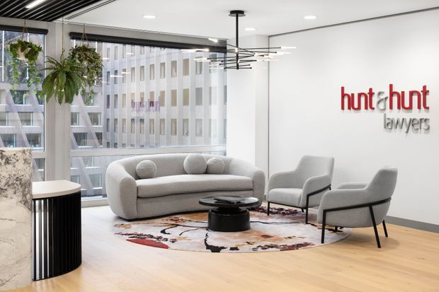 A bright contemporary commercial reception or waiting area for Hunt & Hunt lawyers, featuring a light timber floor, white walls, floor-to-ceiling glazing, a curved pale sofa, two upholstered lounge chairs, a low black coffee table, a patterned area rug, hanging greenery, and a sculptural linear pendant light. A marble-clad reception counter is partially visible at the left.