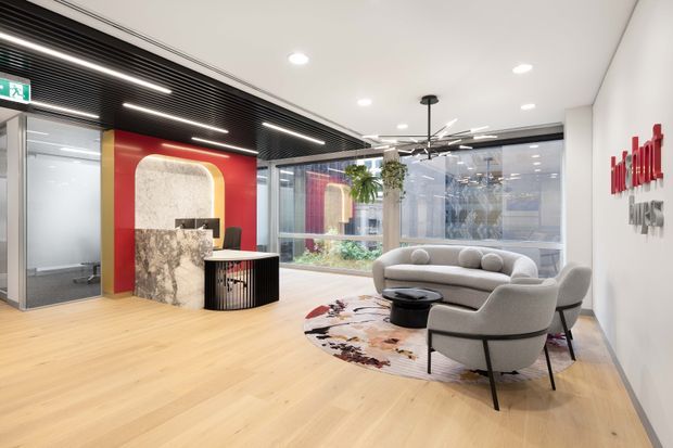 Bright contemporary commercial reception and lounge area with pale timber flooring, a sculptural marble reception desk, bold red feature wall, black slatted ceiling, extensive glazing, and a small seating arrangement around a patterned rug and coffee tables. The space includes a prominent wall-mounted company logo and an adjoining glazed office or meeting room.