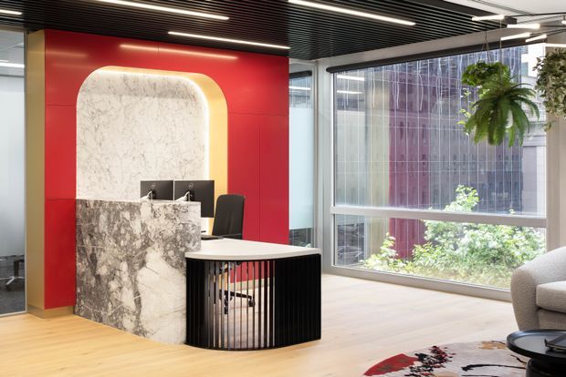 A contemporary commercial reception area with a sculptural marble reception desk set against a vivid red feature wall. The desk incorporates a rounded illuminated stone backdrop and black vertical slatted detailing, while full-height glazing, timber flooring, a black slatted ceiling, indoor plants, and lounge seating complete the polished office interior.