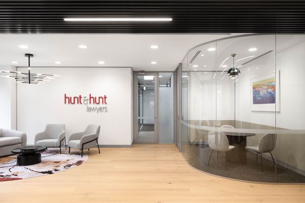 A contemporary commercial law office reception and waiting area. The space features pale timber flooring, white walls, a black linear slatted ceiling with integrated lighting, a minimalist seating arrangement, and a glazed meeting room with a round table and chairs. The firm's logo is displayed on the feature wall.