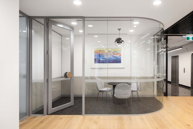 A bright contemporary office interior with a curved frameless glass meeting room enclosed by grey metal-framed glazing. The room contains a round table, upholstered chairs, pendant lighting and wall artwork, with pale timber flooring outside the carpeted meeting space. An adjacent corridor with lift doors is visible on the right.