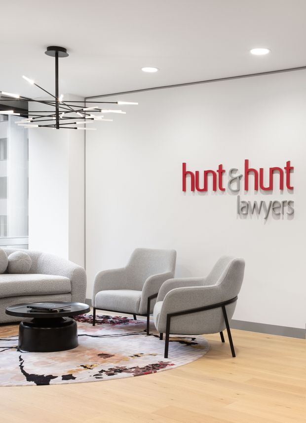 A bright, contemporary law firm reception or waiting area with a minimalist white interior, pale timber flooring, sculptural black-and-white pendant lighting, and a grey upholstered seating arrangement around a black coffee table. A colorful abstract rug anchors the lounge area, while the wall displays the raised 'hunt&hunt lawyers' logo.