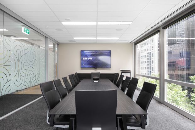 A modern commercial conference room with a long dark wood meeting table surrounded by black upholstered office chairs. The room has floor-to-ceiling windows overlooking an urban setting, a frosted patterned glass partition with a glass door, a wall-mounted display, carpet flooring, and a suspended acoustic tile ceiling. The space is bright, clean, and professional.