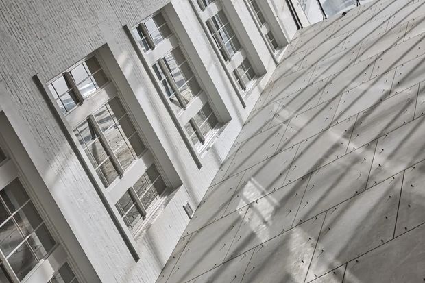 Upward-looking architectural view within a narrow courtyard or atrium, showing tall white-painted brick walls with repeated multi-pane windows and a contrasting sloped or faceted concrete surface. Strong diagonal composition, monochromatic palette, and shadow patterns create a dramatic contemporary institutional or commercial feel.