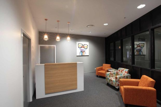 A modern commercial reception or waiting area with a white and timber reception desk, dark framed glazed partitioning, grey carpet, orange lounge chairs, a floral upholstered armchair, pendant lights, and colourful artwork. The space has a clean, contemporary office fit-out with a neutral palette and orange accents.