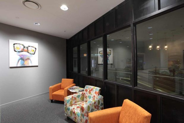 A contemporary commercial waiting or reception lounge with orange upholstered armchairs and a colorful floral-patterned accent chair arranged around a small glass coffee table. The space has dark timber-toned glazed partition joinery, grey walls, grey carpet, recessed ceiling lighting, and artwork on the wall. Reflections through the glazing reveal additional interior areas.
