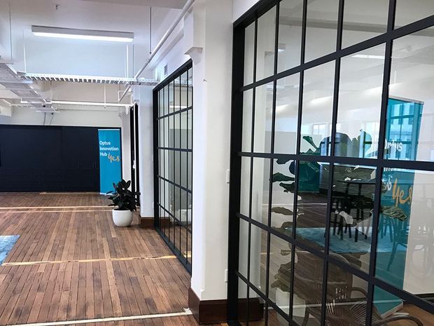 Interior view of a contemporary commercial office or innovation hub with black-framed steel-look glazed partitions, exposed white services and ceiling structure, polished timber flooring, and brightly coloured branded glazing visible through the meeting spaces.