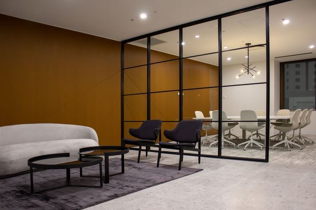 Contemporary office reception and lounge area with a curved light sofa, dark upholstered lounge chairs, nested round coffee tables, and a large grey rug. Black-framed glass partitions separate the lounge from a meeting room furnished with a long table and white rolling office chairs. Warm wood wall panelling, recessed ceiling lighting, and a sculptural pendant create a refined professional interior.