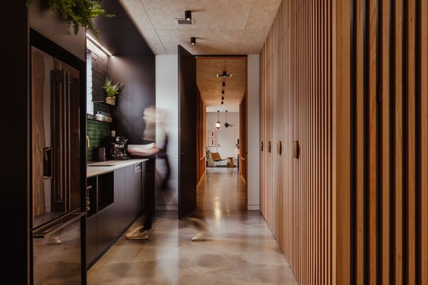 A long, narrow contemporary hallway with dark built-in cabinetry on the left and a full-height vertical timber-slat wall and concealed storage on the right. The corridor has polished concrete or stone-look flooring, a textured light ceiling with a linear arrangement of recessed and pendant lighting, and warm timber door frames leading toward a living area. Black cabinetry, bronze-toned hardware, indoor plants, and warm directional lighting create a refined, moody transition space.