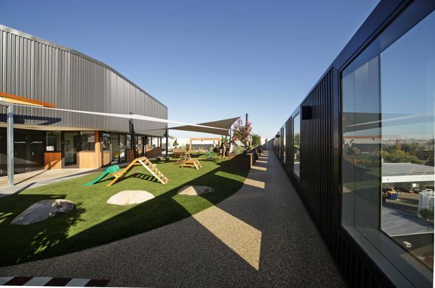 Exterior view of a contemporary commercial or educational complex arranged around a landscaped courtyard. The image shows a dark metal-clad building with extensive reflective glazing on the right, a curved corrugated-metal facade and glazed entrance on the left, and a paved pedestrian walkway beside an artificial-turf recreation area with shade sails and play equipment. The architecture is clean-lined and functional, photographed in bright daylight.
