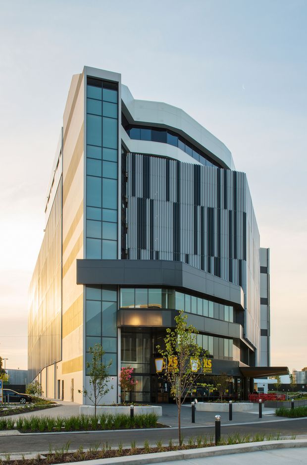 Contemporary multi-storey commercial building photographed from the street, featuring a rounded corner facade, extensive blue-tinted glazing, dark metal framing, vertical metal sun-shading fins, and a light-colored solid side wall. The ground level contains a glazed commercial entrance and illuminated signage, with landscaped planting and pedestrian paving in the foreground.