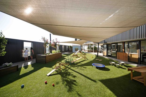 A landscaped outdoor courtyard or terrace enclosed by low dark-clad contemporary buildings. Large stretched shade sails cover an artificial-grass recreation area with outdoor seating, planter boxes, a small trampoline, and children’s play equipment. The surrounding buildings feature black vertical cladding, extensive glazing, and sheltered walkways, creating a bright communal outdoor space.