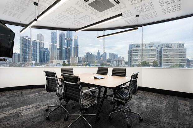 A modern commercial meeting room with a large conference table and black ergonomic office chairs, set against floor-to-ceiling glazing overlooking a dense urban skyline. The room has dark patterned carpet, a white acoustic ceiling with integrated services, and suspended linear pendant lighting.