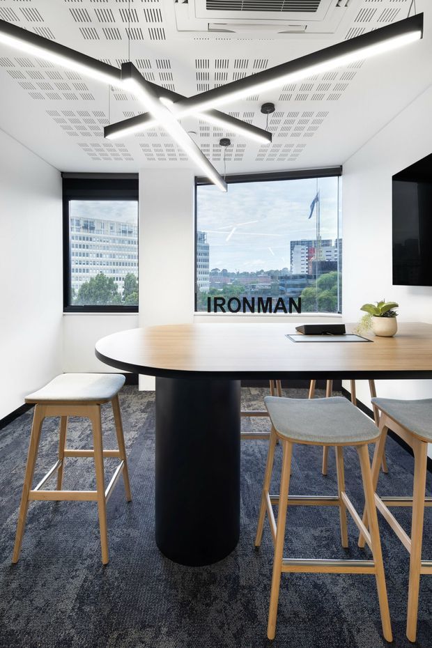 Contemporary commercial breakout or meeting area with a large oval timber table on a black cylindrical pedestal, surrounded by light timber stools with grey upholstered seats. The bright white room has acoustic perforated ceiling panels, striking black linear pendant lighting, dark carpet, roller blinds, and large windows overlooking an urban setting. A wall-mounted screen and potted plant complete the workspace.