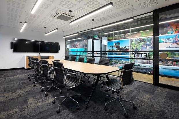 Modern commercial boardroom with a large oval light-wood conference table, black leather swivel chairs, wall-mounted displays, and expansive glazed walls featuring colorful graphic imagery. The room has dark patterned carpet, a perforated acoustic ceiling, linear pendant lighting, and a clean corporate fit-out.