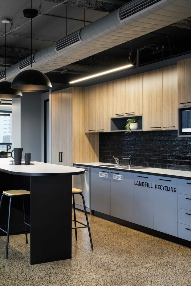 A contemporary commercial kitchenette or staff breakout kitchen with full-height light timber cabinetry, a black glossy brick-tile splashback, pale benchtops, and grey lower cabinets labelled for landfill and recycling. A black island with bar stools sits in the foreground. The ceiling has exposed services, large round HVAC ductwork, black pendant lights, and linear strip lighting. The floor is speckled terrazzo-like concrete.