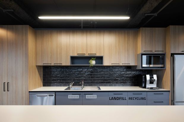 Compact contemporary kitchenette with full-height warm wood-grain cabinetry, matte dark blue-grey lower cabinets, a pale benchtop, glossy black brick-look splashback, stainless sink and appliances. The ceiling is exposed and painted black with linear fluorescent lighting. Recycling and landfill labels are printed on the cabinetry, suggesting a workplace or commercial break-room setting.