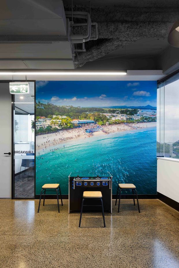 A modern commercial interior featuring a large beach panorama wall graphic, a foosball table with stools, glass partitions, exposed dark services and ductwork, linear LED lighting, and a polished exposed-aggregate concrete floor. An exit door and glazing are visible at the sides.