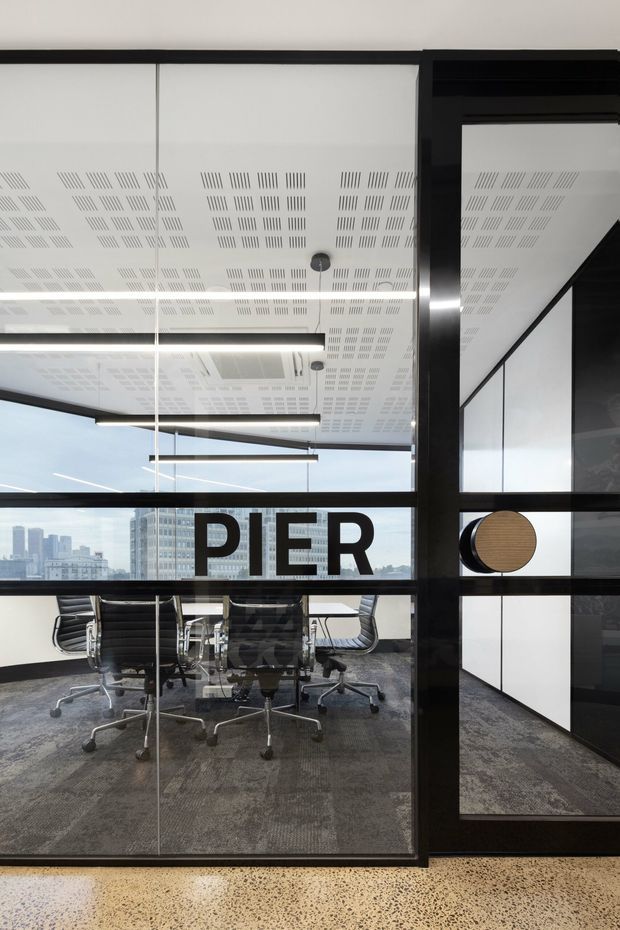A contemporary commercial office meeting room viewed through black-framed glass partitions. The room contains a long conference table surrounded by black office chairs, with a city skyline visible through the glazing. The interior features a perforated acoustic ceiling with integrated linear lighting, dark carpet, black framing and cabinetry, and a polished terrazzo-like floor in the foreground. Large “PIER” lettering is applied to the glass.