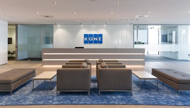 Contemporary corporate reception and waiting area with a central illuminated reception desk, KONE branding, glass-partitioned offices, neutral upholstered lounge seating, white wall paneling, and a large blue patterned carpet.