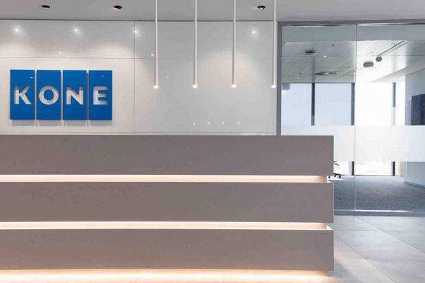 Modern commercial reception area featuring a large minimalist desk with illuminated horizontal fronts, a white glossy wall behind it, hanging linear pendant lights, and a prominent KONE logo. Glazed partitions and an open office or lobby area are visible to the right, with a combination of carpet and large-format tile flooring.