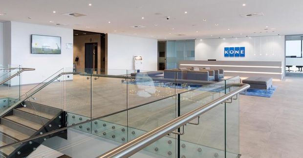 Bright contemporary commercial reception or lobby with a prominent glazed stair and stainless-steel handrail in the foreground. The space features pale stone or concrete-look flooring, white walls and ceiling, glass balustrades, a blue-grey lounge seating area, and a branded reception backdrop.