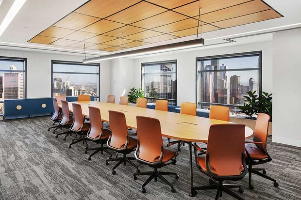 Bright contemporary commercial meeting room with a long light-wood conference table surrounded by orange and tan swivel chairs. Floor-to-ceiling corner glazing provides city views, while a geometric timber-panel ceiling feature, suspended linear lighting, patterned carpet, indoor plants, and blue built-in storage complete the space.