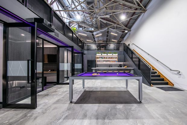 A contemporary commercial-style recreational space with an exposed steel-truss ceiling, polished concrete floor, dark glazed partitions and joinery, a purple-lit pool table, a bar and beverage display at the rear, and a staircase with timber handrail along the right wall. The space has an industrial, high-contrast fit-out with bright overhead lighting.