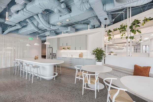 A bright contemporary commercial breakout and kitchen area with a long white island bench and bar stools, pale built-in cabinetry, integrated appliances, pendant lights, café tables and chairs, indoor plants, and extensive exposed white-painted HVAC ductwork overhead. The space has polished concrete or terrazzo-style flooring and a glazed, open-plan office character.