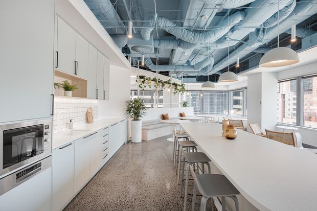 Bright contemporary open-plan office breakout and kitchen area with white full-height cabinetry, integrated appliances, a long white island with bar seating, built-in bench seating, indoor plants, pendant lighting, and exposed services across the ceiling. Large windows provide an urban outlook.