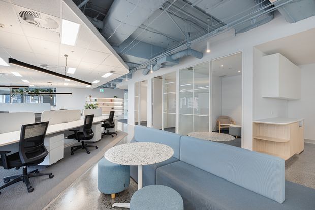 Bright contemporary open-plan office interior with exposed services and ductwork, glazed meeting rooms, workstation desks, ergonomic office chairs, and a pale blue lounge seating area with round tables. The space has polished concrete flooring, white built-in cabinetry, suspended lighting, and a clean professional fit-out.