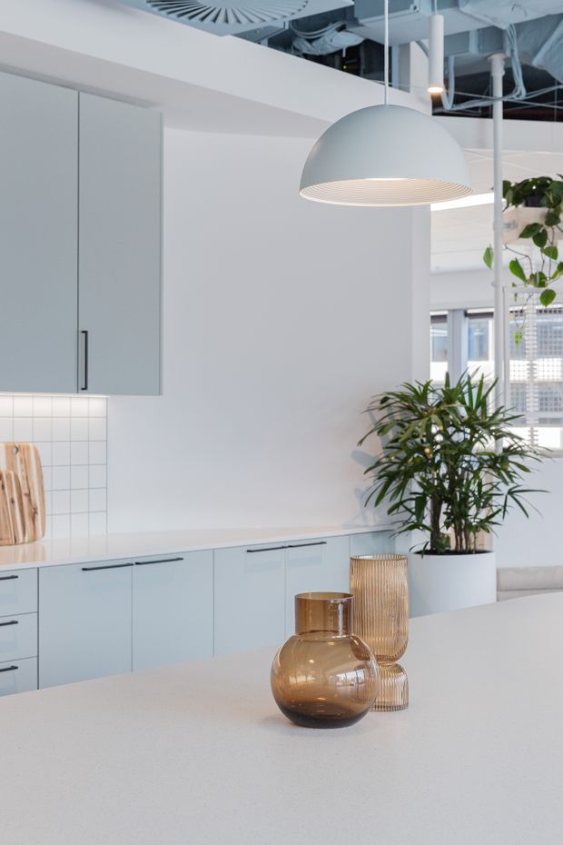 Bright, minimalist kitchen interior with pale blue-grey flat-panel cabinetry, a light stone island benchtop, white tiled splashback, and a large sculptural pendant light. Exposed services at the ceiling and abundant indoor greenery add an industrial-commercial character, while amber glass vessels provide warm accents.
