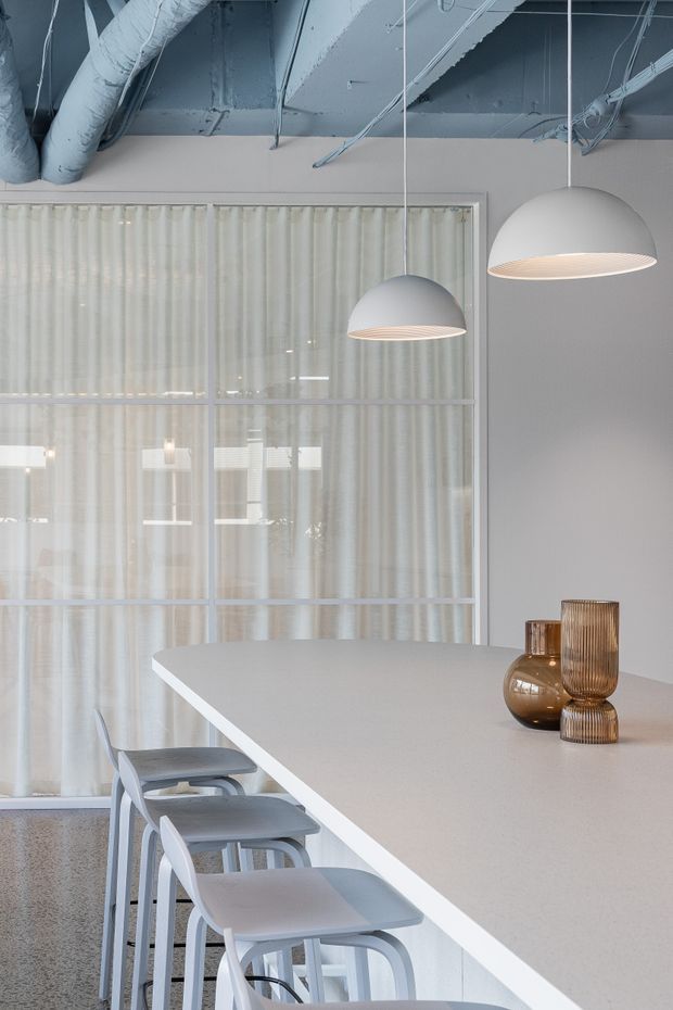 A bright contemporary kitchen or kitchenette with a long white island and integrated seating, sheer-curtained glazed partition, exposed painted services and ductwork, and two large white pendant lights. The restrained palette of white, pale grey, and translucent bronze-toned glass accessories creates a clean, refined atmosphere.