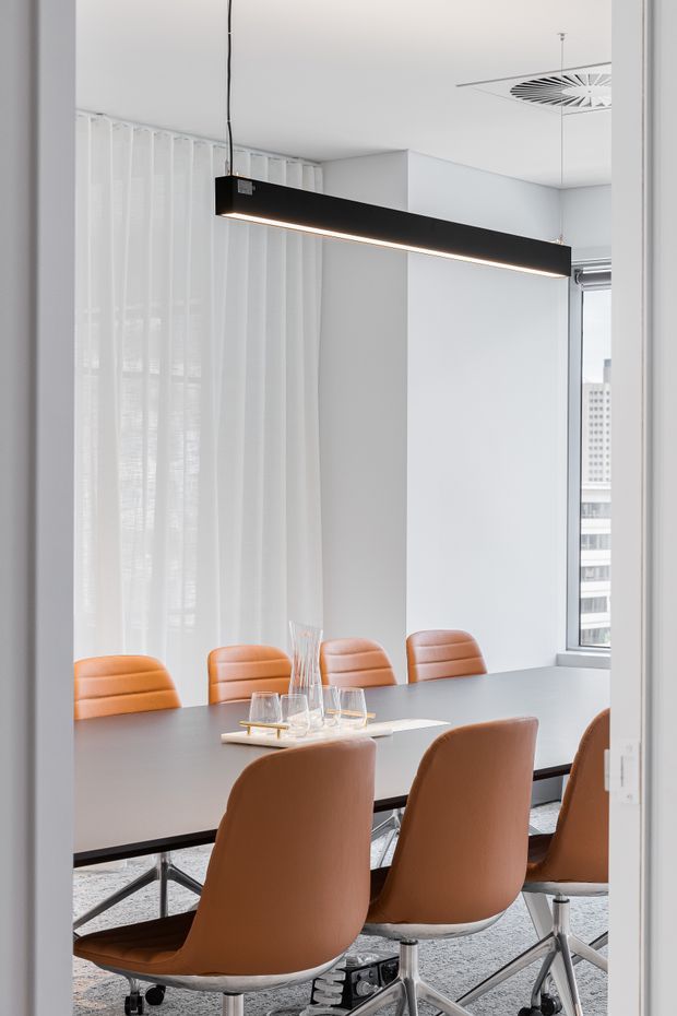 Bright contemporary dining or boardroom-style meeting space with a long dark rectangular table, multiple caramel leather swivel chairs, sheer white curtains, white walls, a linear suspended black pendant light, and a modern ceiling air diffuser. High-rise city buildings are visible through the window.