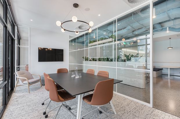 Contemporary office meeting room with a large dark rectangular conference table, warm tan swivel chairs, a sculptural circular pendant light, wall-mounted screen, and floor-to-ceiling glazing. The glass partitions reveal an open-plan workspace with exposed services, polished concrete-style flooring, and integrated planter boxes.
