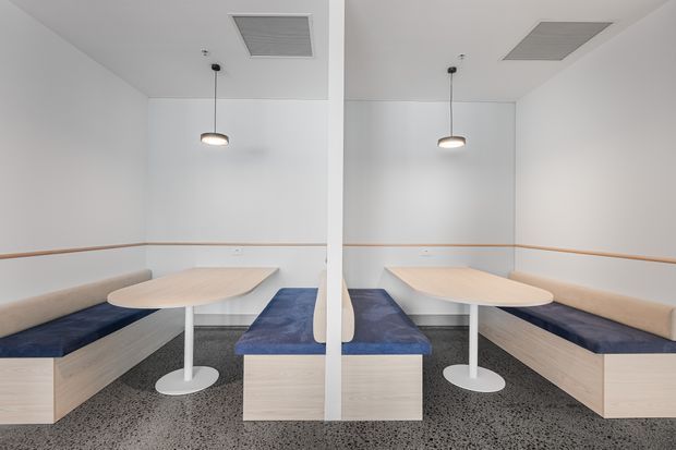 A bright, minimalist built-in seating area with two mirrored booth configurations divided by a central partition. Pale wood bench bases and tabletops are paired with deep blue upholstered cushions, white pedestal tables, white walls, slim timber wall rails, pendant lights, and a grey speckled floor.