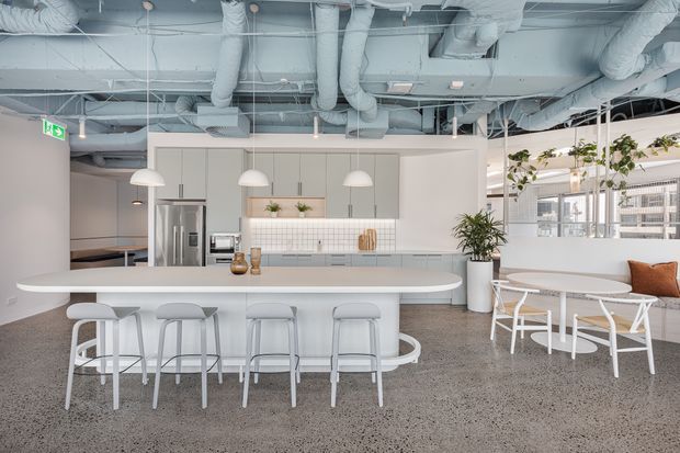 Bright open-plan commercial breakout and kitchen area with a large white island bench, integrated seating, pendant lights, pale cabinetry, tiled splashback, exposed services ceiling, and a casual lounge beside full-height glazing. The space has a clean contemporary workplace fit-out with polished concrete flooring and indoor plants.