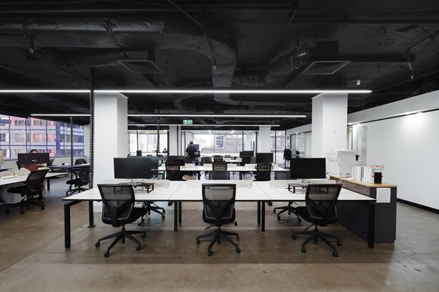 Open-plan commercial office with shared white desks, black ergonomic task chairs, computer monitors, and workstations arranged across a polished concrete floor. The space features exposed black ceiling services and ductwork, white structural columns, extensive glazing, and a large white wall, creating a contemporary industrial workplace setting.